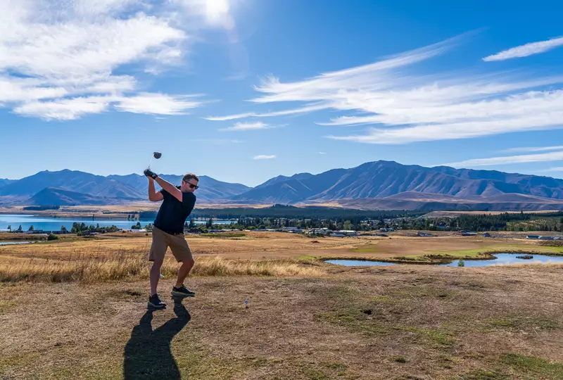 Image of a man, swinging gold club set to tee off on a course in Mackenzie District overlooking lakes and mountains